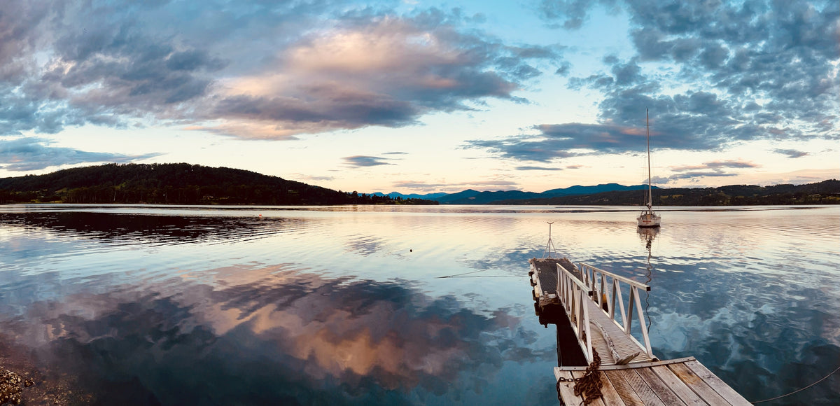 A scenic lake landscape at sunset or sunrise with dramatic clouds reflected in calm water, a wooden dock extending into the lake, a small sailboat moored near the right side, and dark tree-covered hills in the distance.